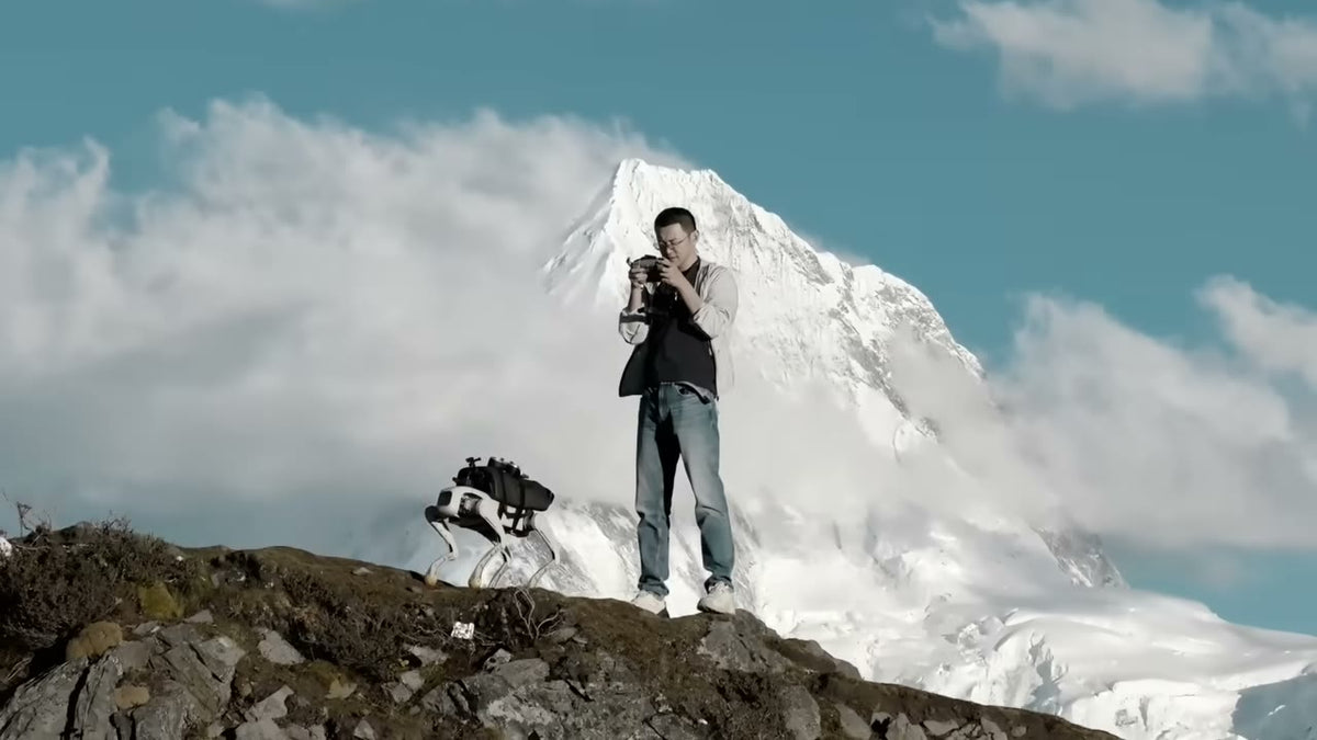 Operator with an AgiBot D1 Pro on a rocky summit in front of a snowy mountain